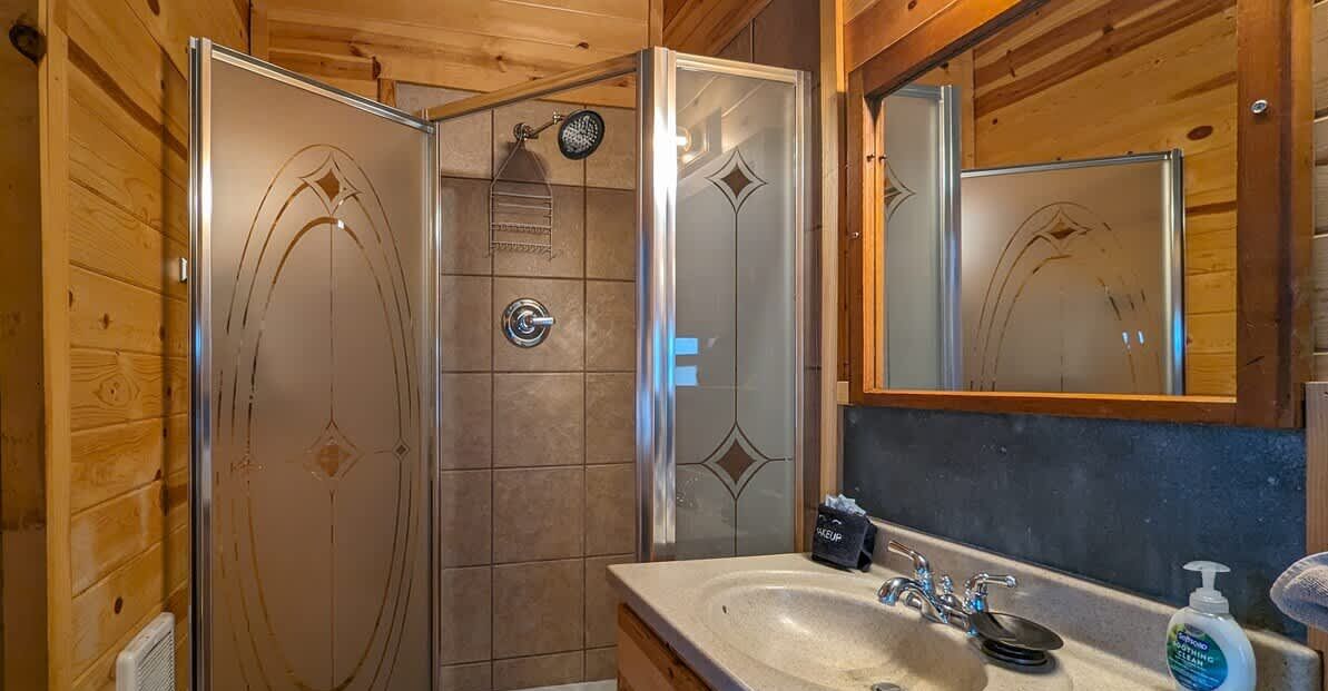 Baño de cabaña de madera con mampara de vidrio, lavamanos con espejo y un perchero para toallas.