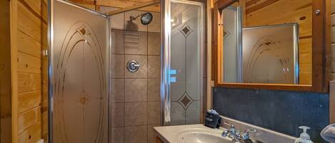 Baño de cabaña de madera con mampara de vidrio, lavamanos con espejo y un perchero para toallas.
