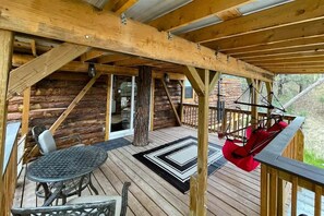 Una terraza de madera con una mesa y sillas, un sillón colgante y vistas al interior de una cabaña de troncos.