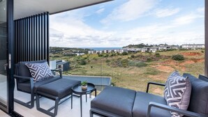A balcony with a modern sofa and chairs, a small table with a plant, and a view of a residential area and hills. - 1 Savili - Zululami (Ballito)