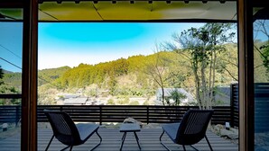 A wooden deck with two black chairs and a small table, overlooking a forested hillside with houses in the background. - Tagomori no Akari (Ukiha)
