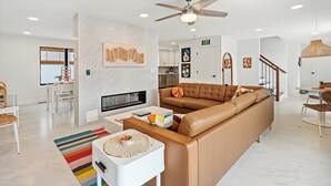 A modern living room with a leather sofa, a coffee table, and a fireplace. There is a dining area with a table and chairs, and a kitchen in the background.
