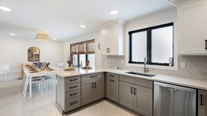 A modern kitchen with white cabinetry, a stainless steel dishwasher, and a dining area with a white table and chairs.