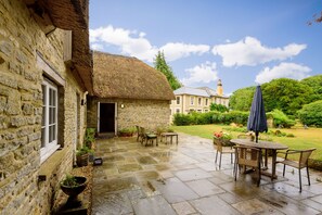 Guests can dine or relax on the patio, surrounded by a well-maintained garden and a charming stone building.