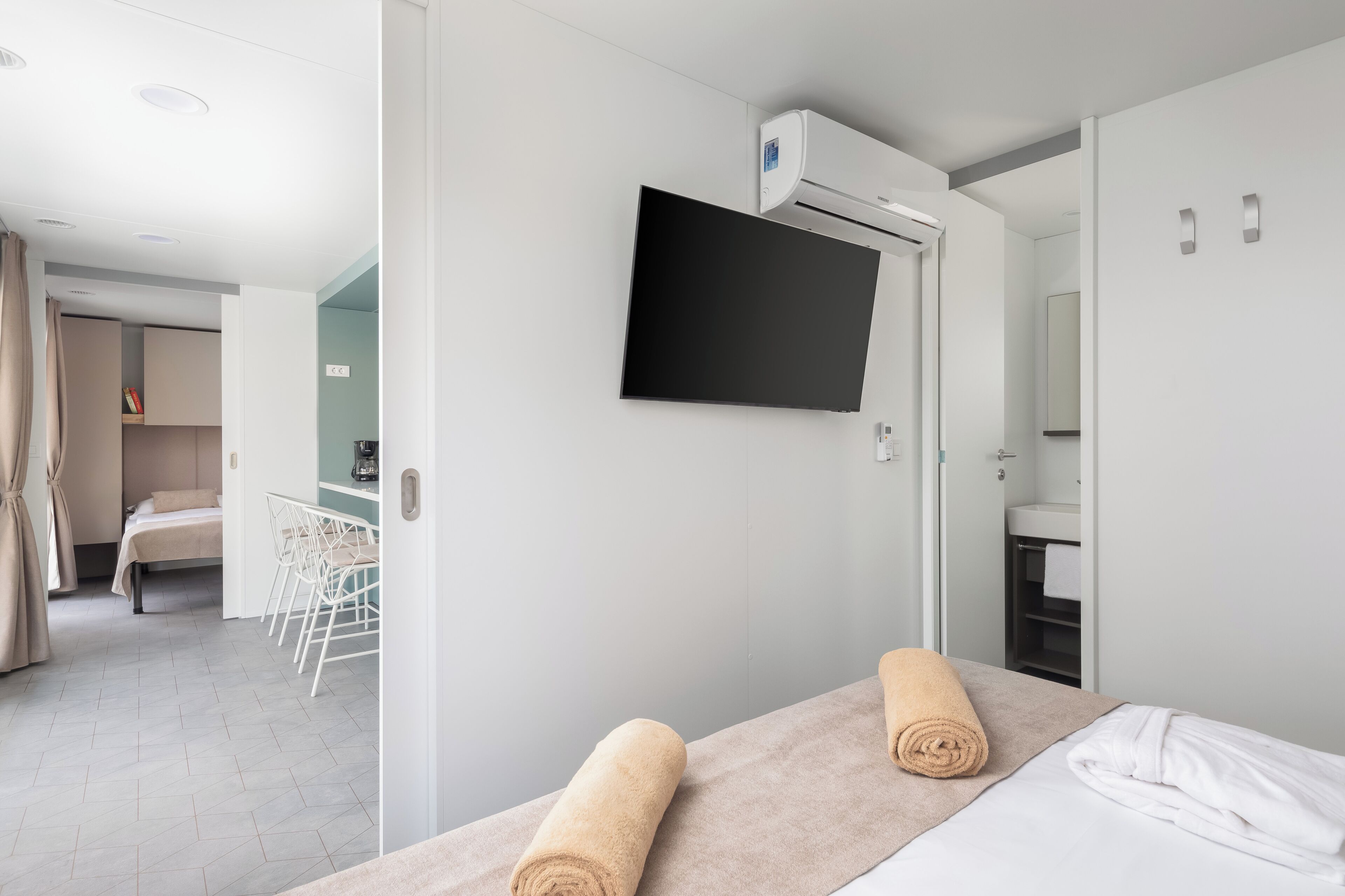 A hotel room with a bed, a TV mounted on the wall, a bathroom with a sink, and a small seating area with chairs.