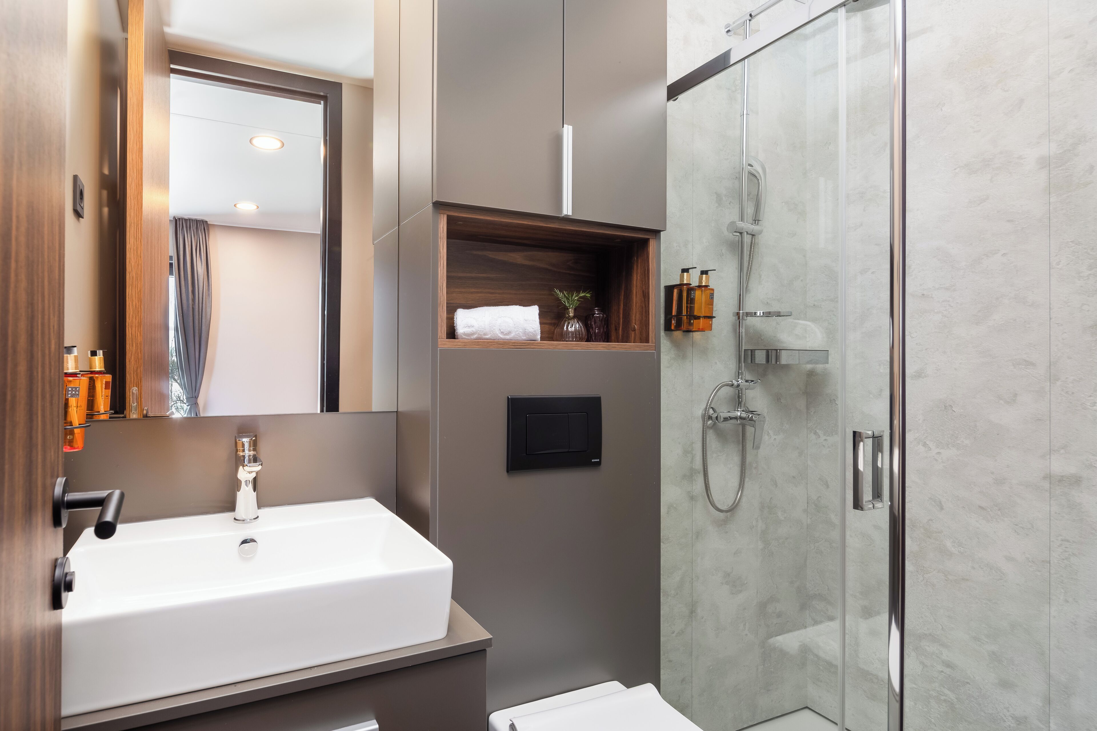 A modern bathroom with a large white sink, a glass shower enclosure, and a wooden shelf with toiletries.