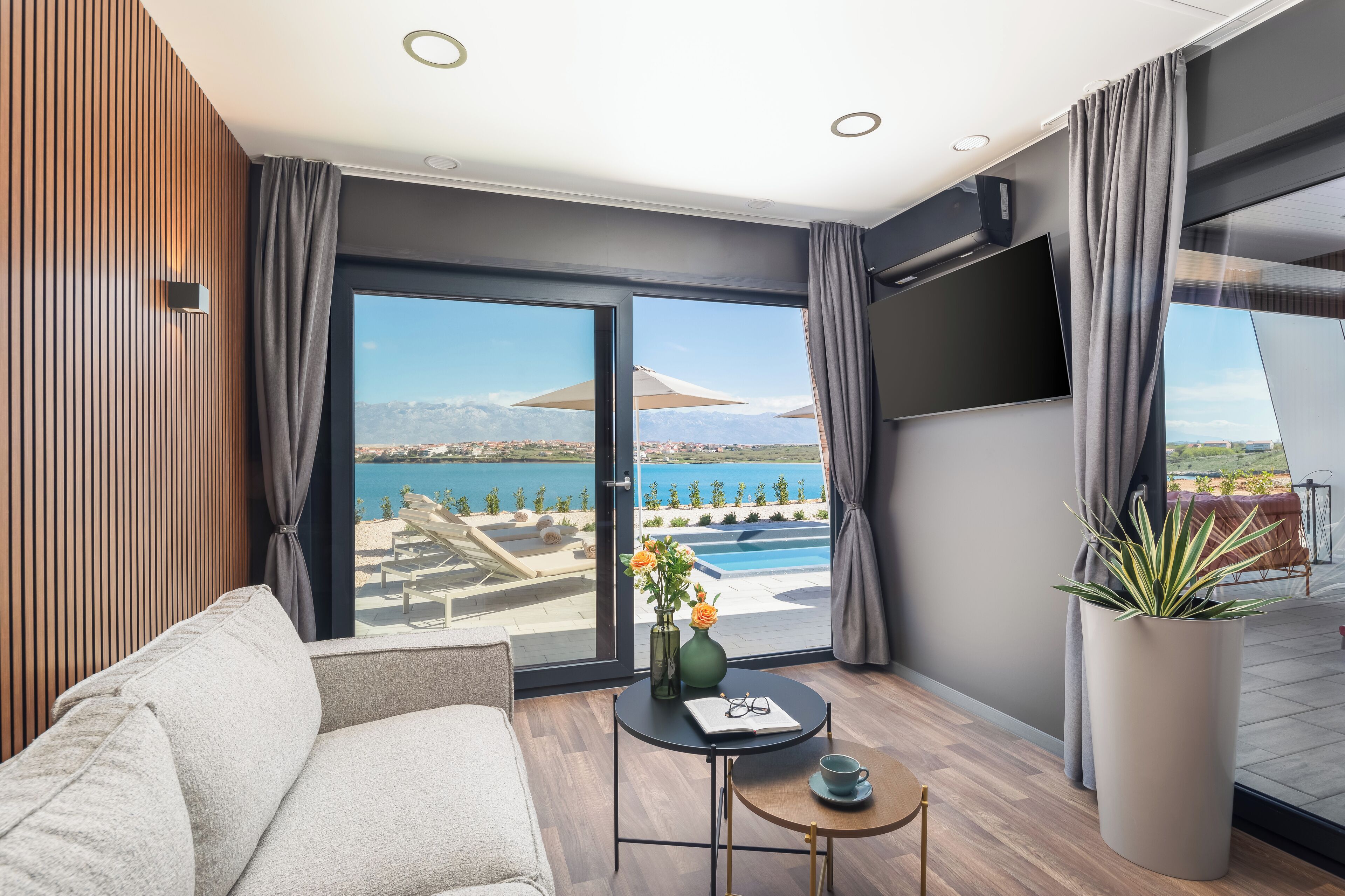 Ein modernes Wohnzimmer mit einer Couch, einem kleinen Tisch mit Blumenvase und Blick auf ein Schwimmbad und Berge.