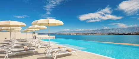 Ein Poolbereich mit Liegestühlen und Sonnenschirmen mit Blick auf ein Gewässer und Berge.