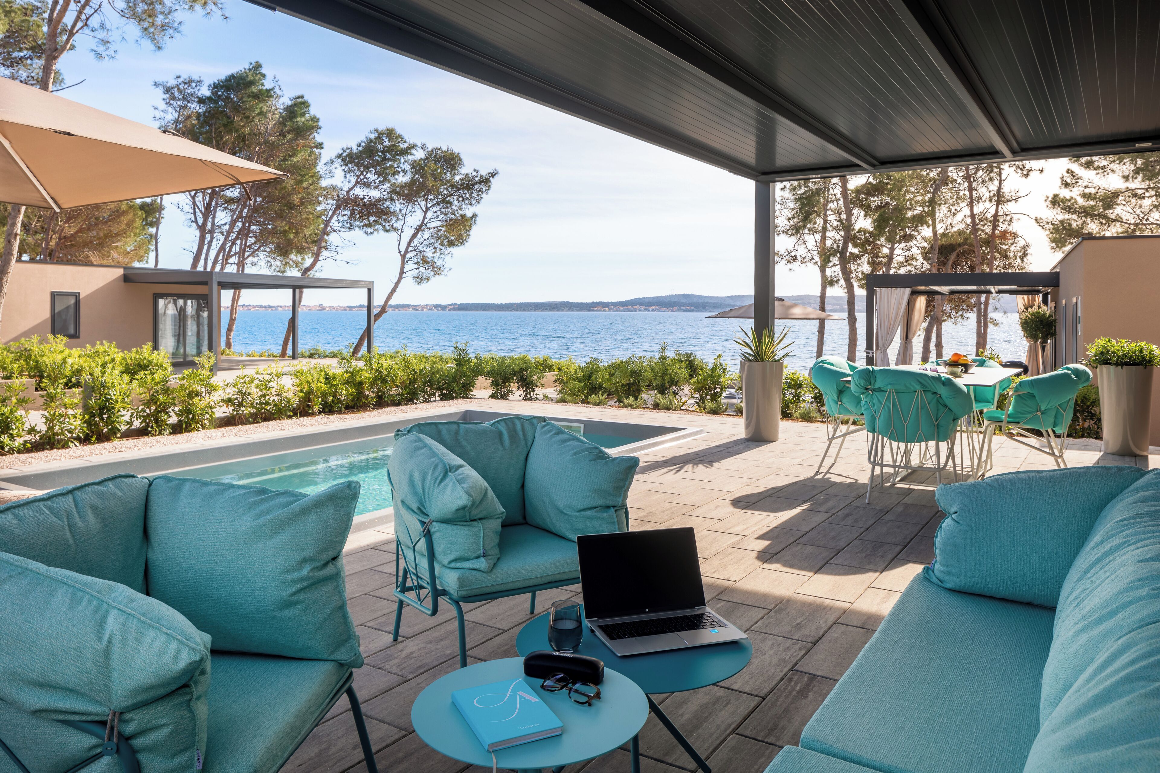 A modern outdoor seating area with a laptop, a notebook, and glasses on a table, overlooking a pool and the sea.