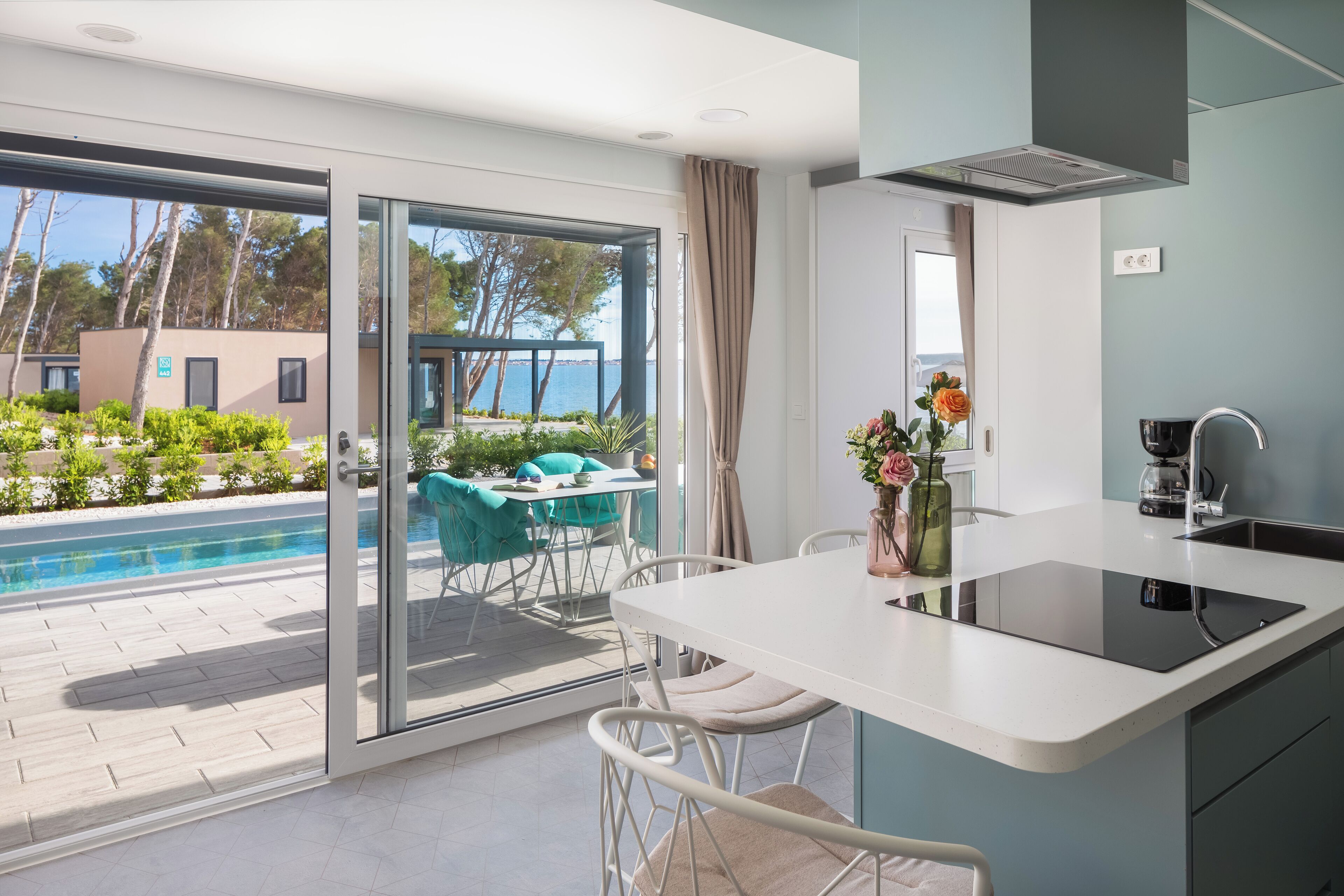 Una cocina moderna con una isla grande, un comedor con sillas y vistas a una piscina y árboles.