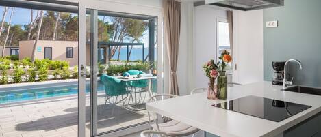 Una cocina moderna con una isla grande, un comedor con sillas y vistas a una piscina y árboles.