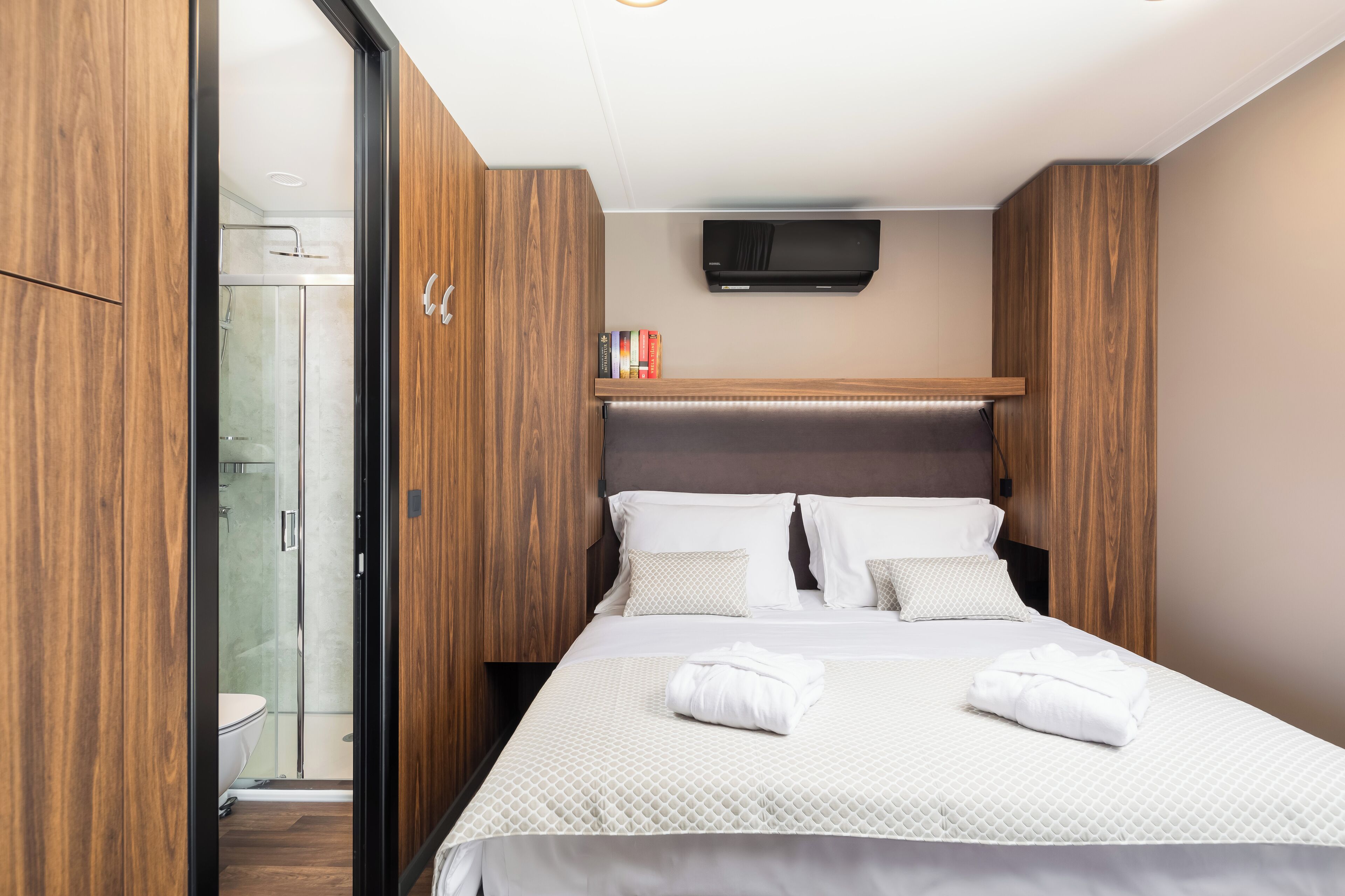 A hotel room with a bed, a television mounted above it, a shower area, and wooden paneling on the walls.