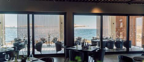 An indoor dining area with tables and chairs, large windows with a view of the sea, and a wooden ceiling.
