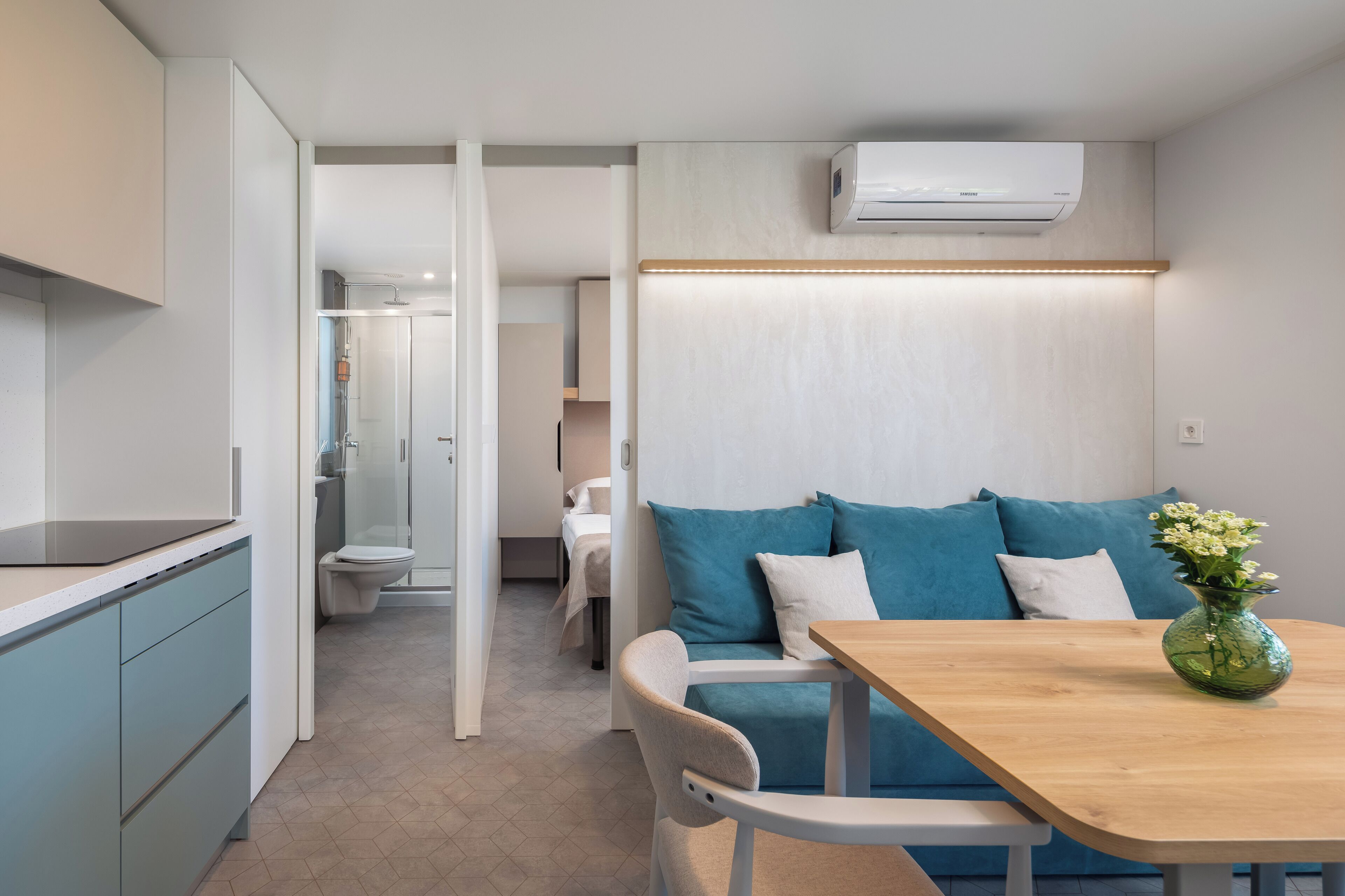 Un espace de vie moderne et compact, comprenant un coin cuisine, un espace repas avec une table en bois et un canapé agrémenté de coussins bleus.