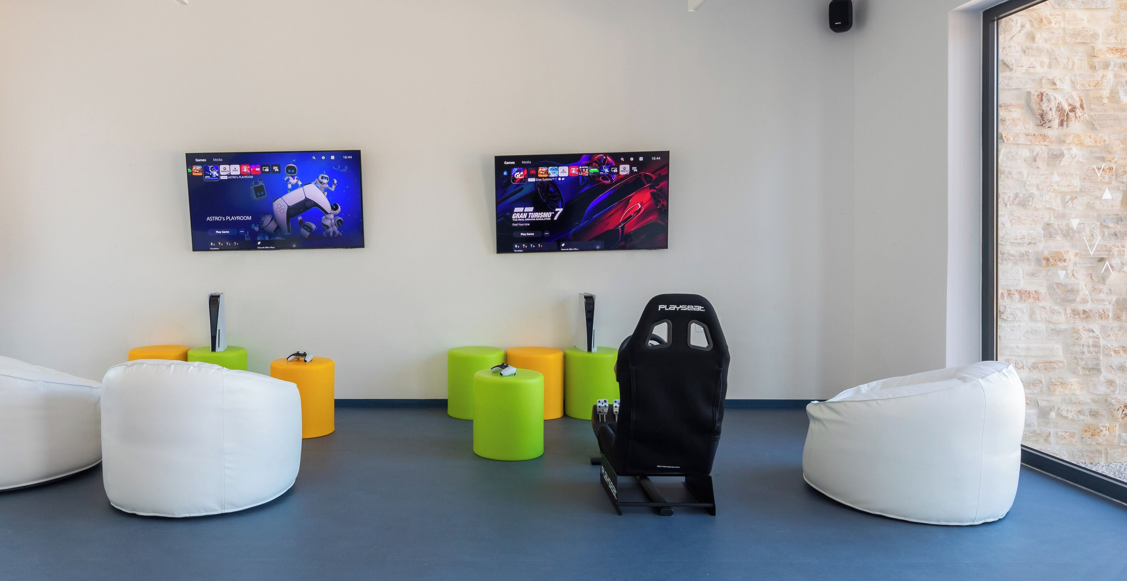 Ein Gaming-Raum mit zwei Monitoren, die Spiele zeigen, einem Playseat-Gaming-Sessel und bunten Sitzsäcken.