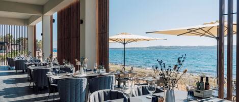 An outdoor dining area with tables and chairs, large umbrellas, and a view of the sea.