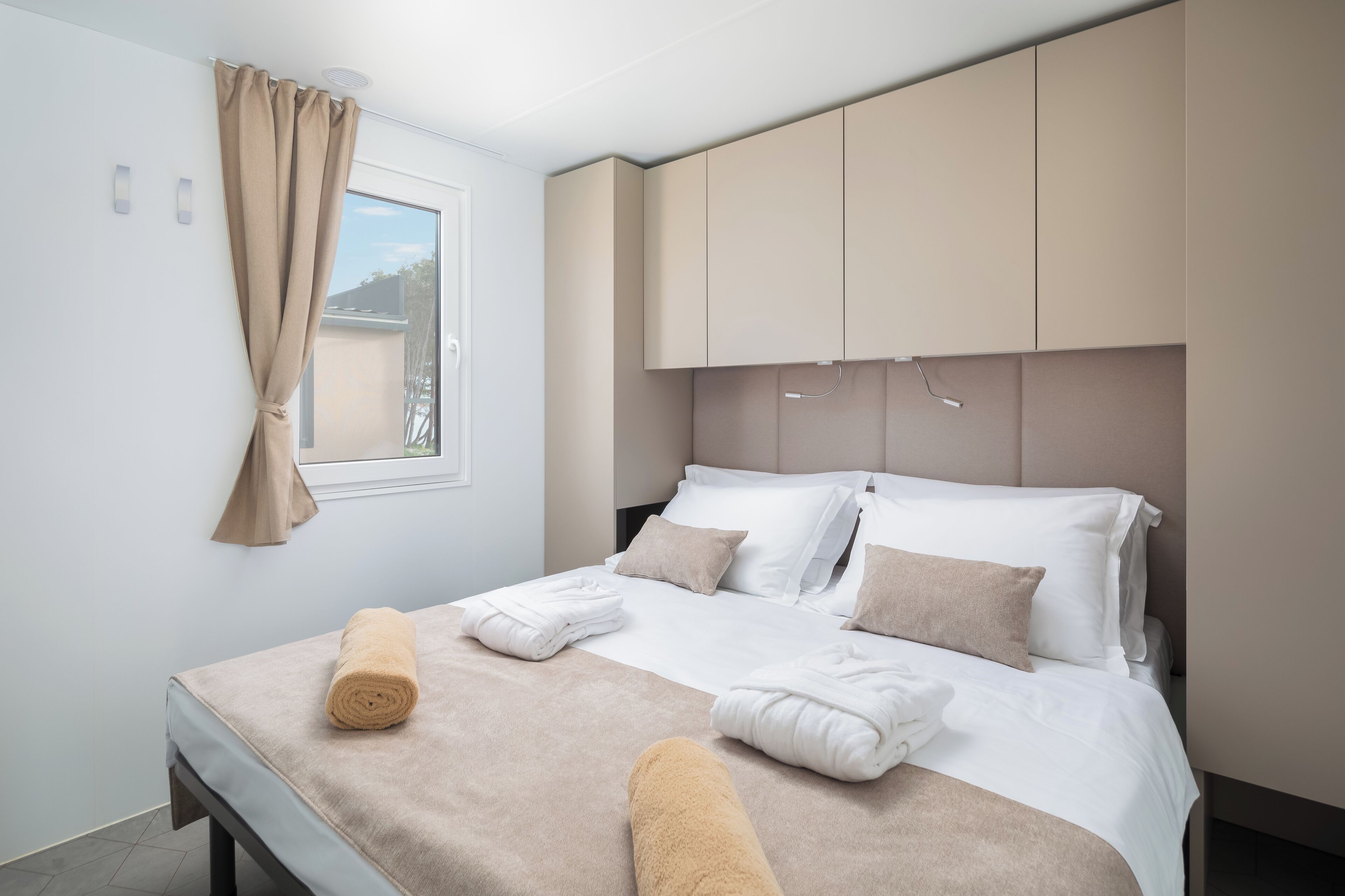 Una cama bien hecha con ropa de cama blanca y beige, una ventana con cortinas beige y un cabecero con iluminación integrada.