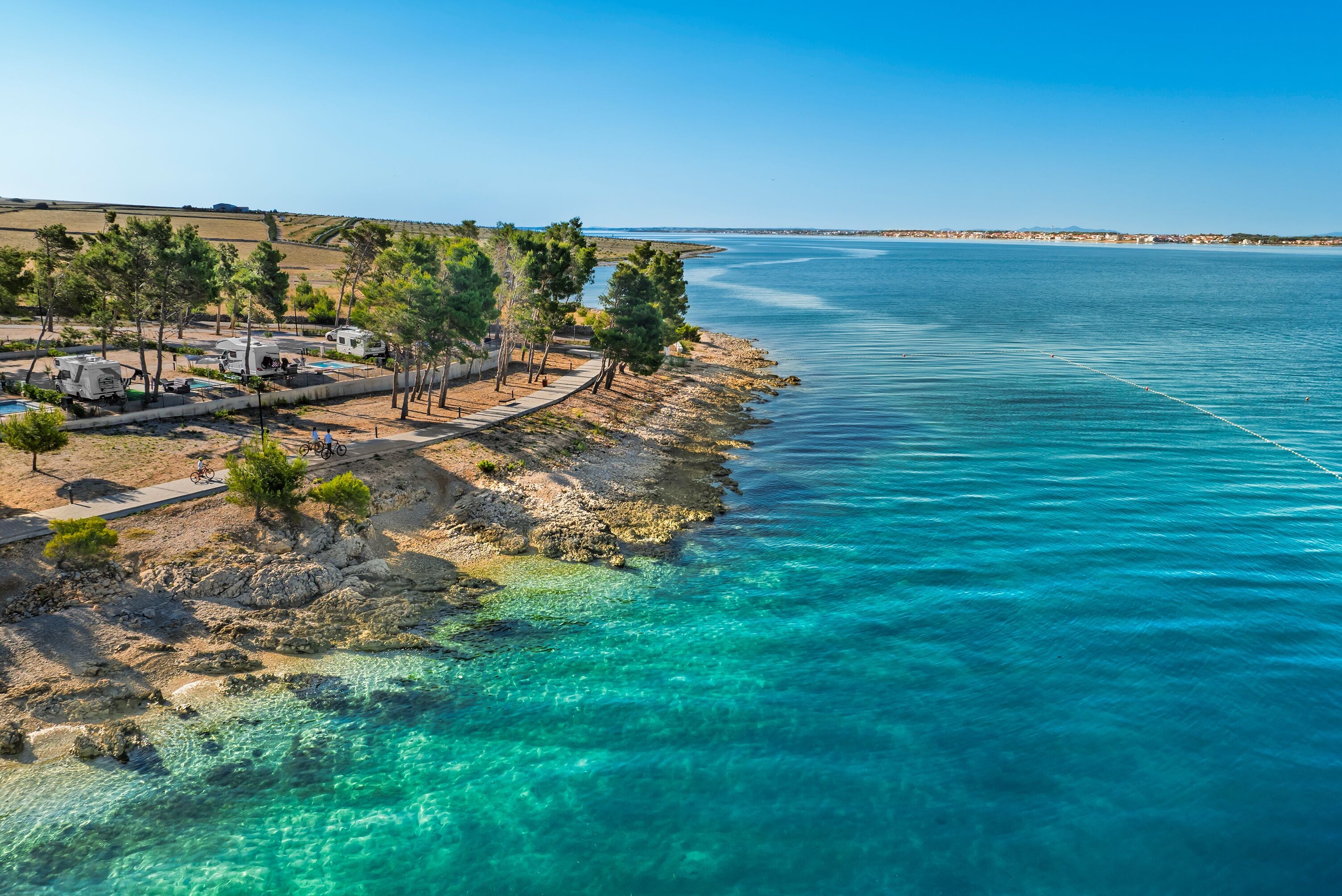 A coastal area with RVs parked along a rocky shoreline, clear turquoise water, and a distant view of a town.