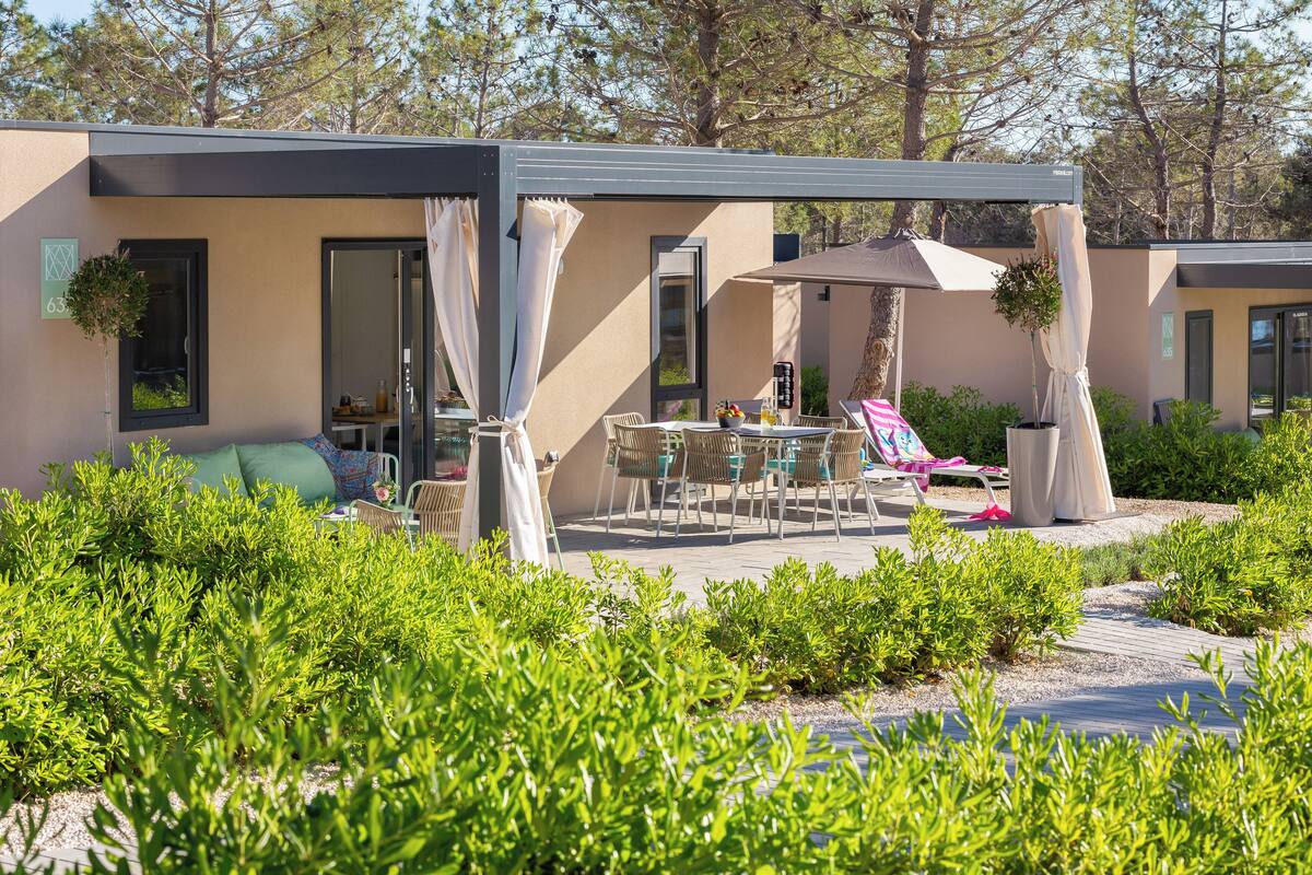 Ein moderner Außenbereich mit Terrasse, Tisch und Stühlen, umgeben von viel Grün und einem überdachten Sitzbereich.