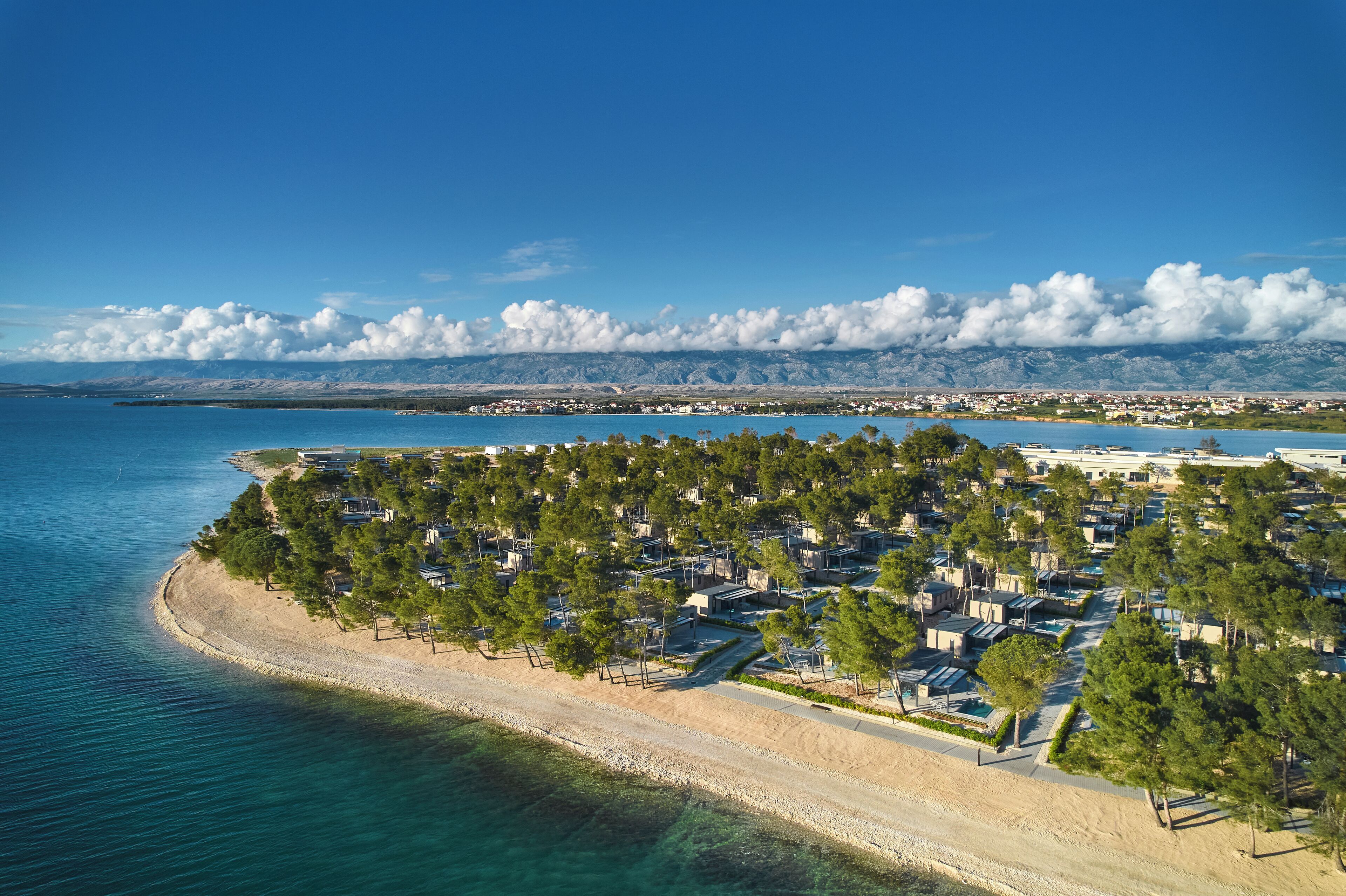 An aerial view of a coastal resort with a sandy beach, dense greenery, and a clear blue sky.
