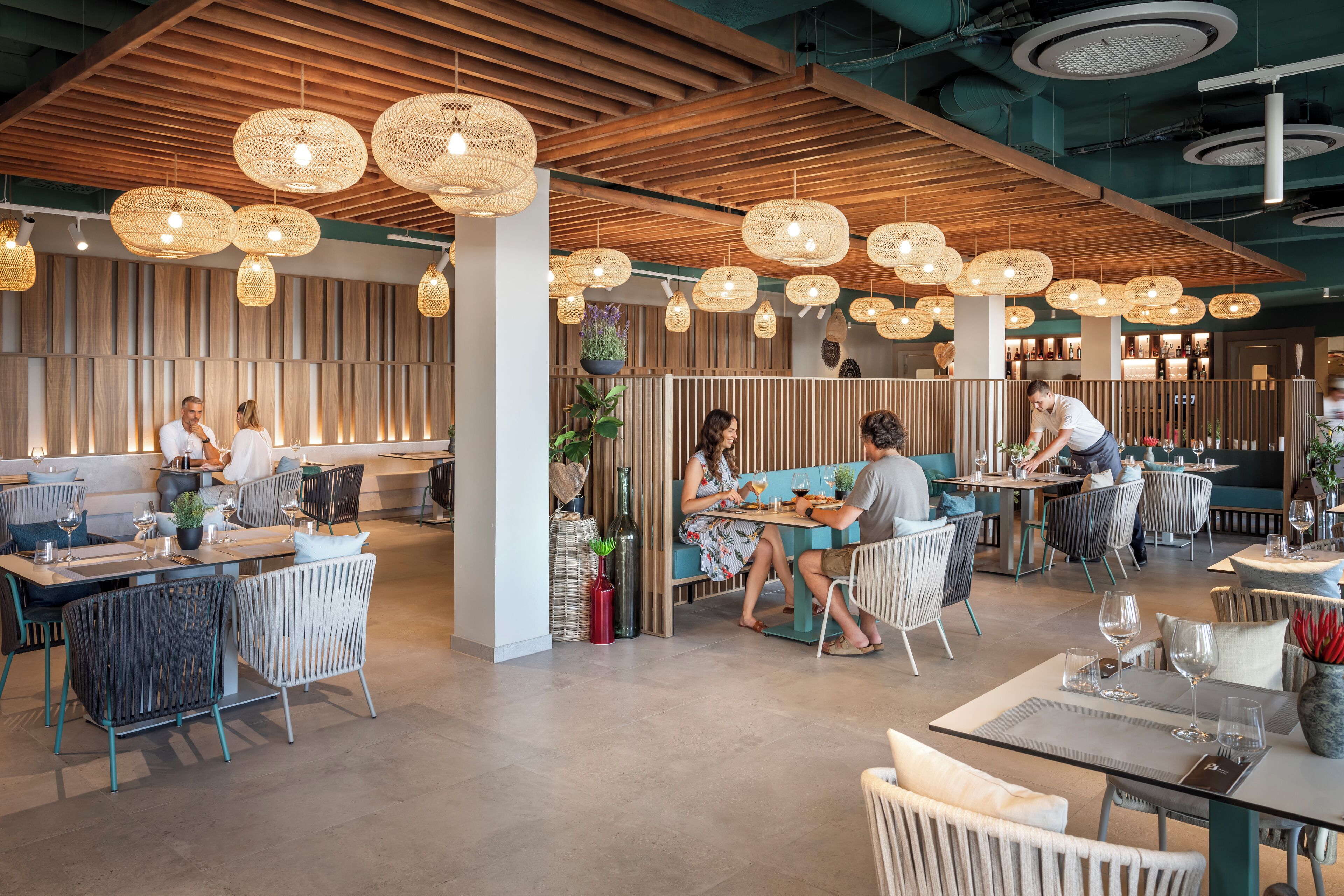 Un restaurante moderno con vigas de techo de madera, lámparas colgantes tejidas y una variedad de arreglos de asientos, incluyendo mesas y cabinas.