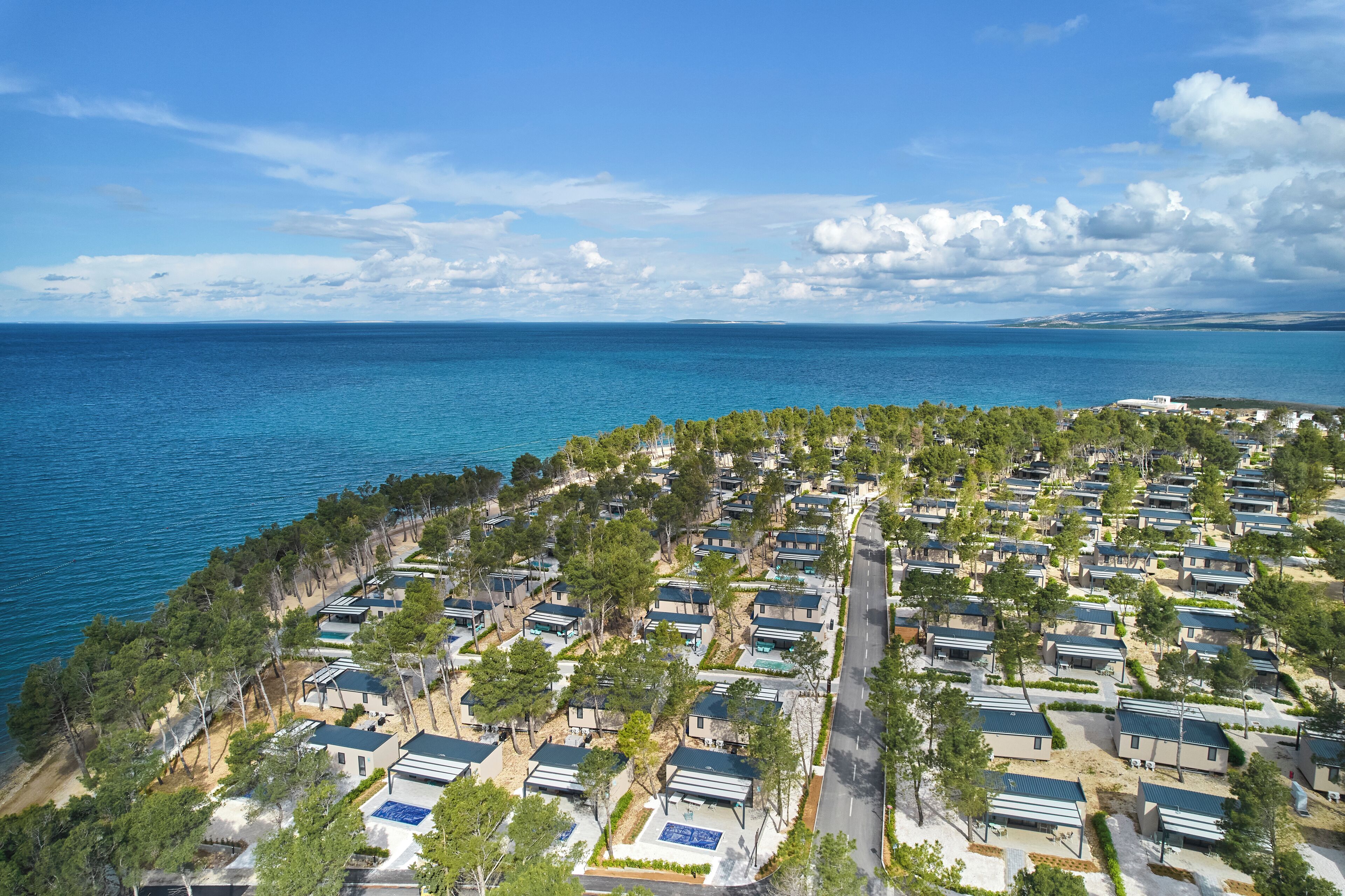 An aerial view of a coastal resort with rows of cabins, a clear blue sea, and a distant landmass.