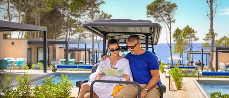 Una pareja en un carrito de golf cerca de una piscina con vistas al mar y a árboles.