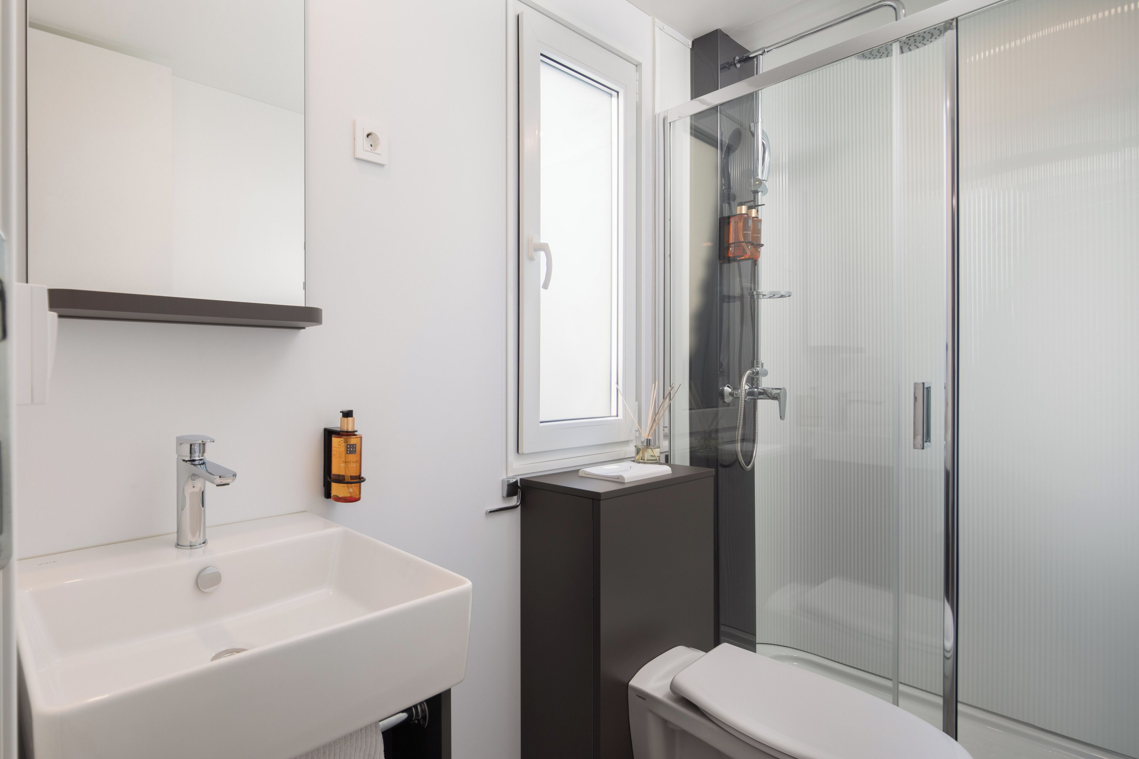 Un bagno moderno con un lavandino bianco, uno specchio, cabina doccia in vetro e un water.