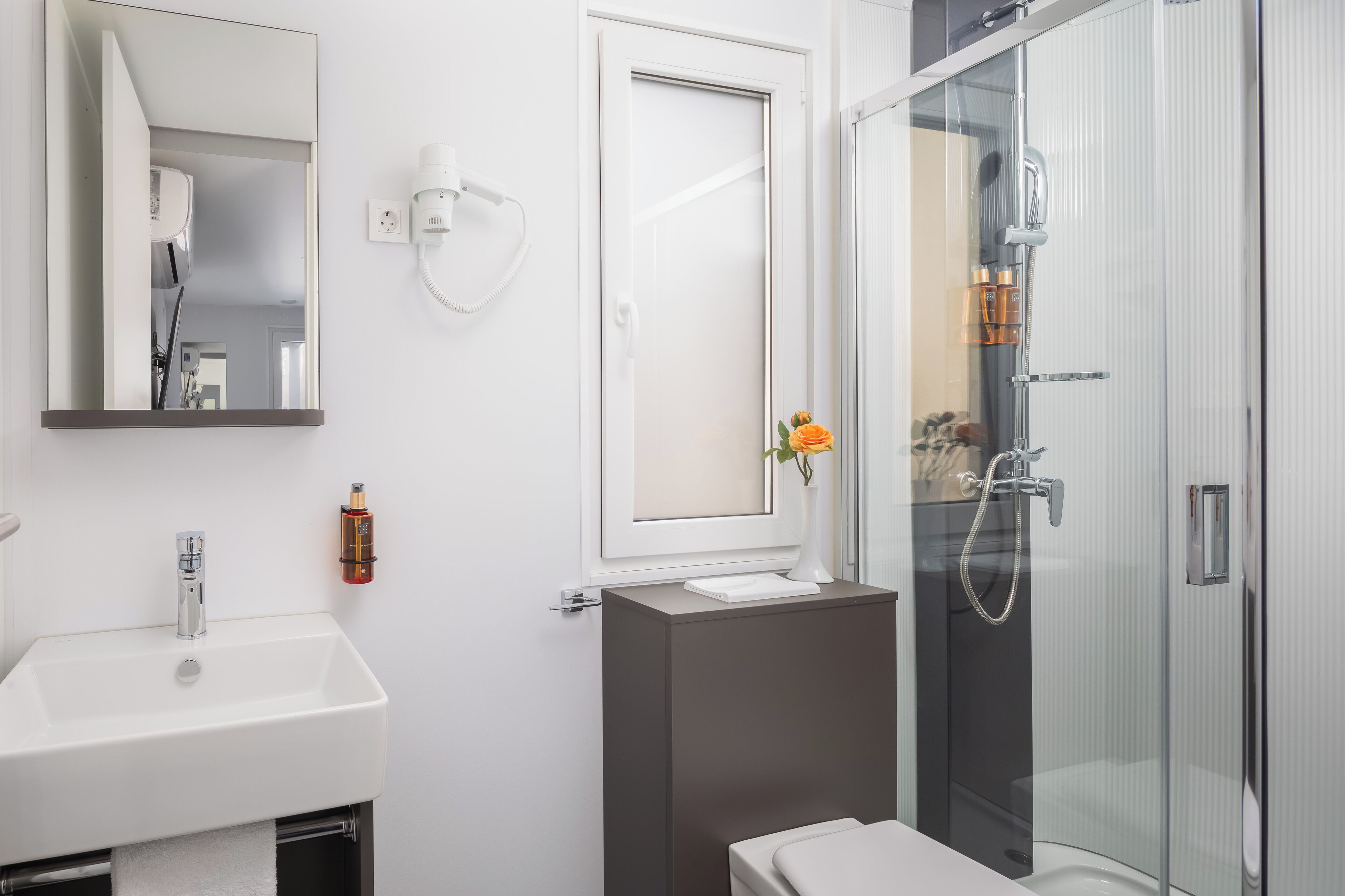 A modern bathroom with a white sink, a mirror, a glass shower enclosure, and a small vase with flowers.