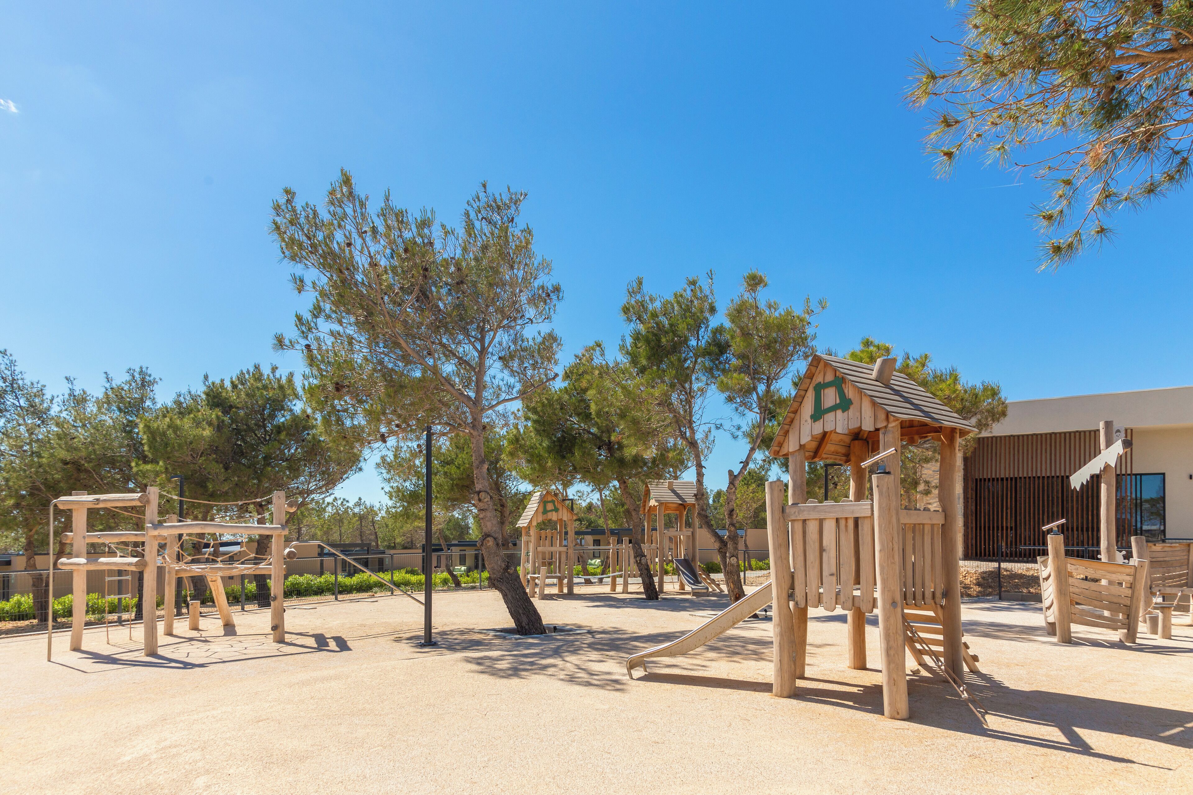 A playground with wooden climbing structures and slides, surrounded by trees and a building in the background.