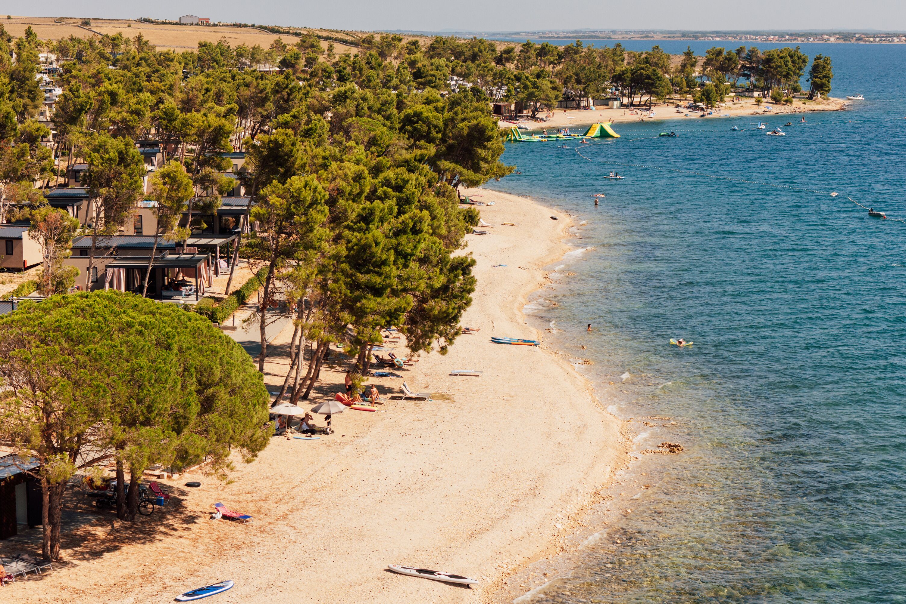 A beach with clear blue water, a sandy shore, and several boats. There are trees and a few buildings in the background.