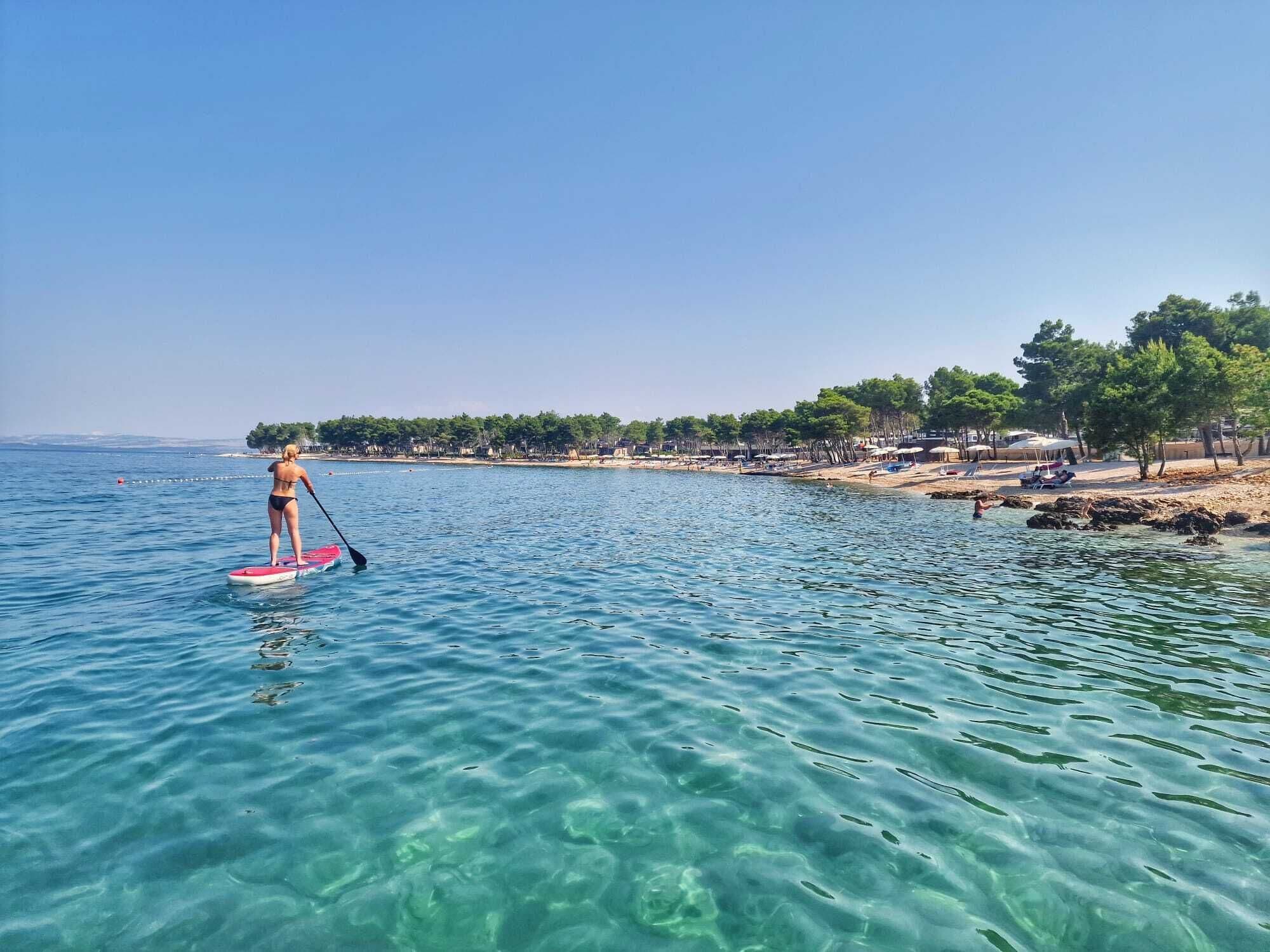 A beach scene with clear water, a paddleboarder, and a beach with umbrellas and trees.