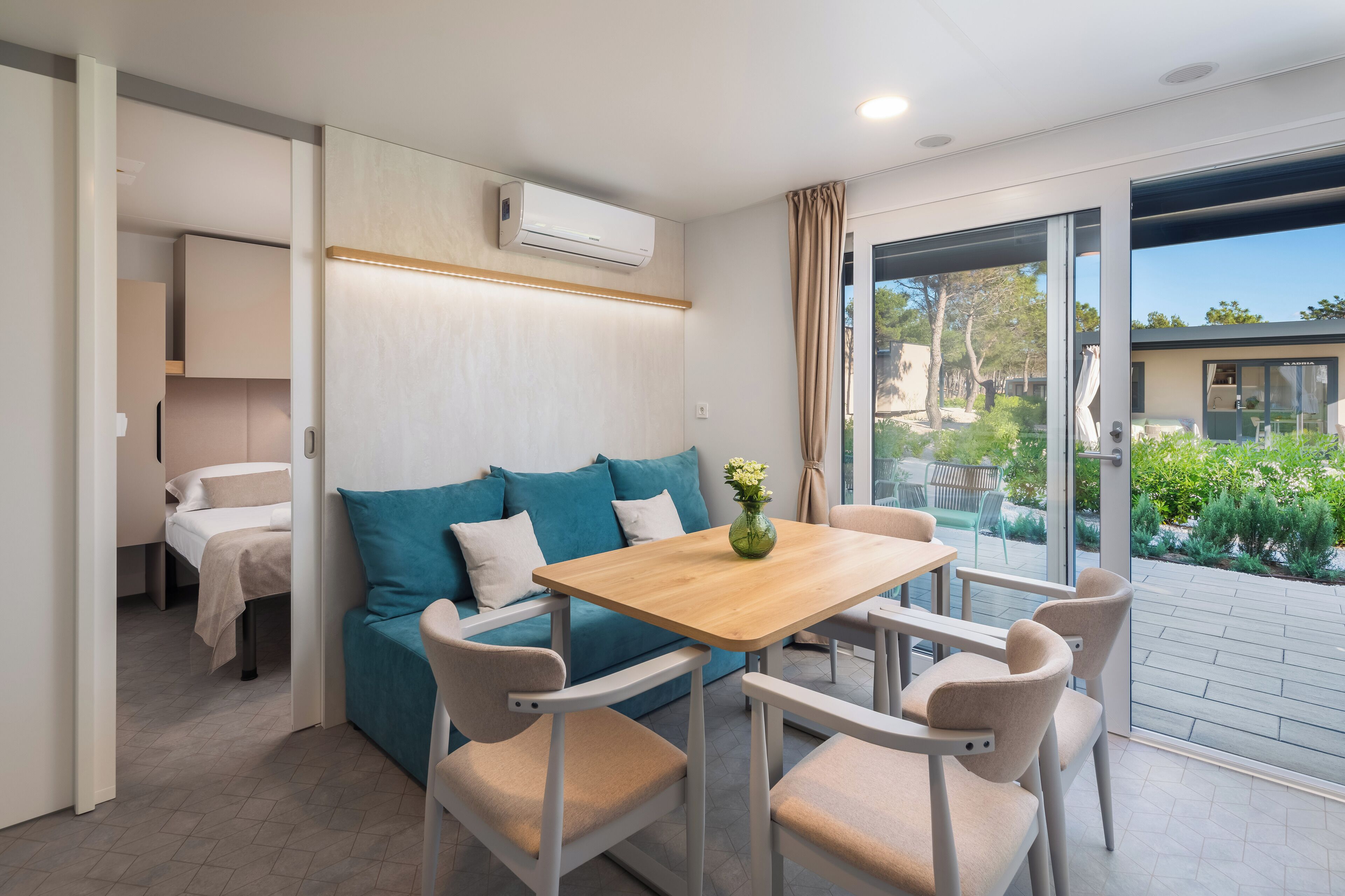 Una habitación moderna con una mesa de madera, sillas color beige, un sofá azul y un ventanal de vidrio que da a un espacio exterior.