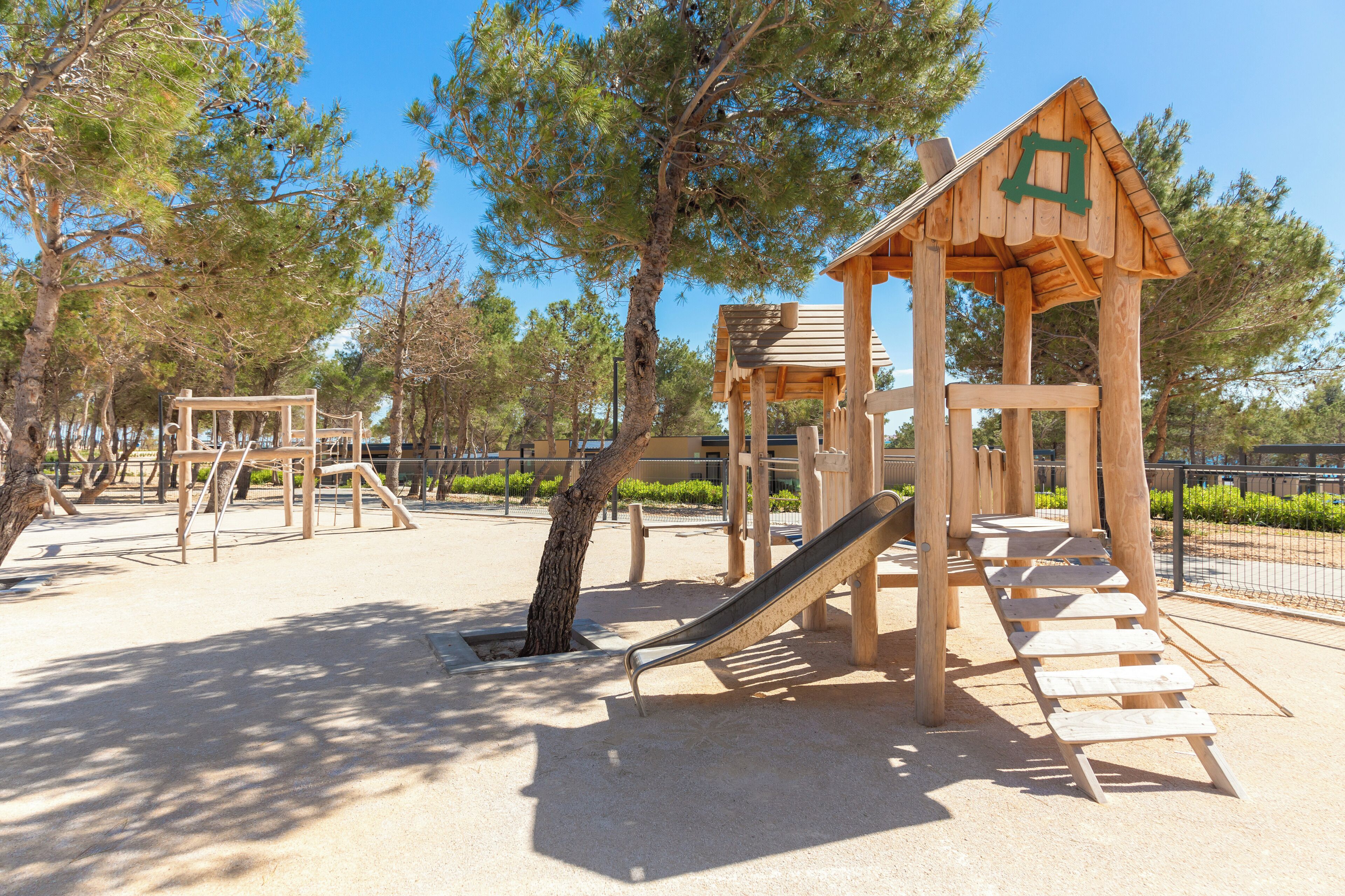 Ein Spielplatz mit Holzkonstruktionen, darunter eine Rutsche und Klettergerüste, umgeben von Bäumen.