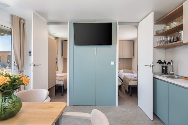 A compact hotel room with a kitchenette, a flat-screen TV mounted on a wall, a dining area with a table and chairs, and a bathroom with a shower curtain. - Aminess Style Camping Avalona Glamping Villas & Holiday Homes (Povljana)