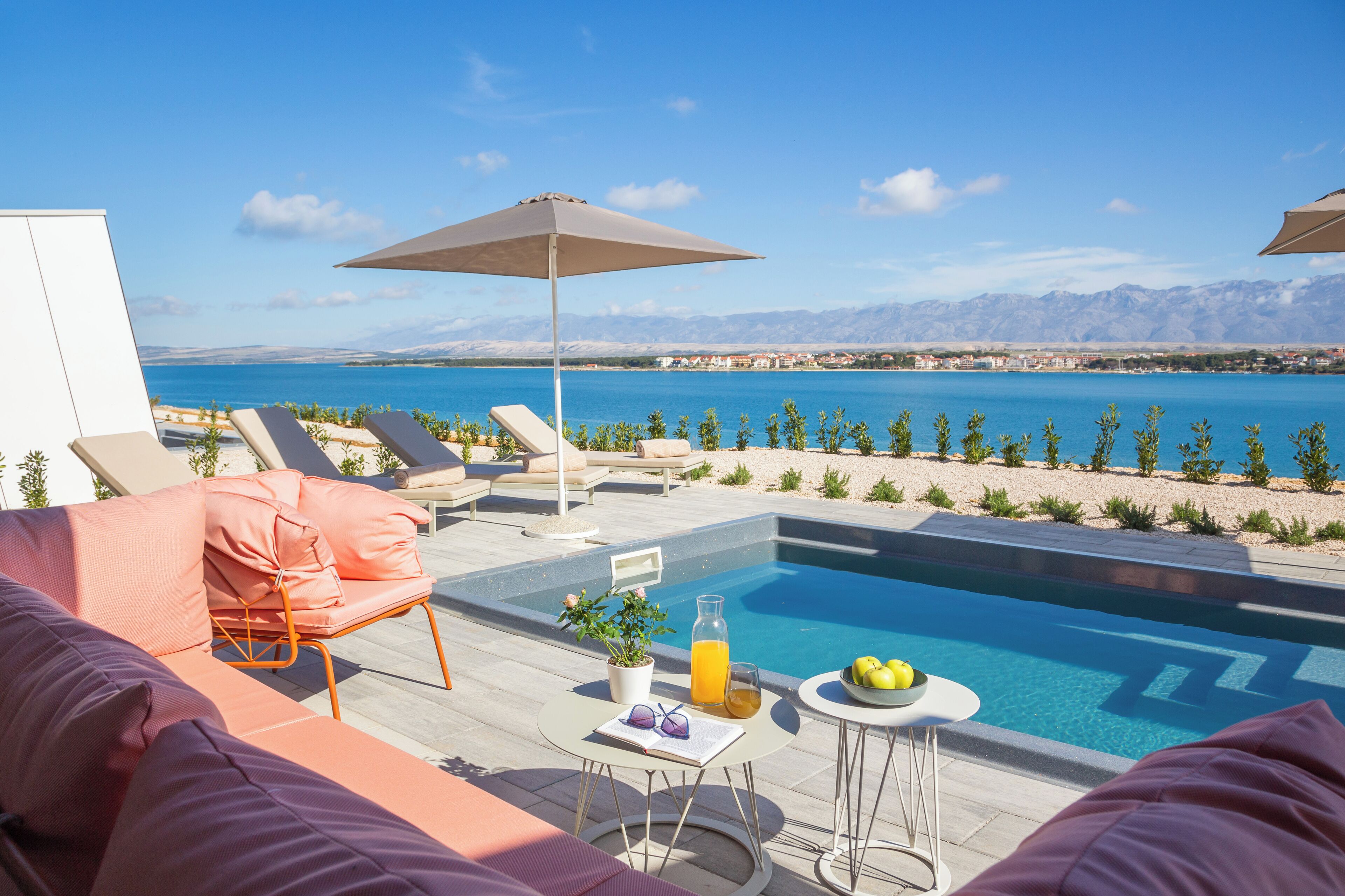 Zona piscina con lettini, un tavolo con un drink e vista sul mare e sulle montagne.