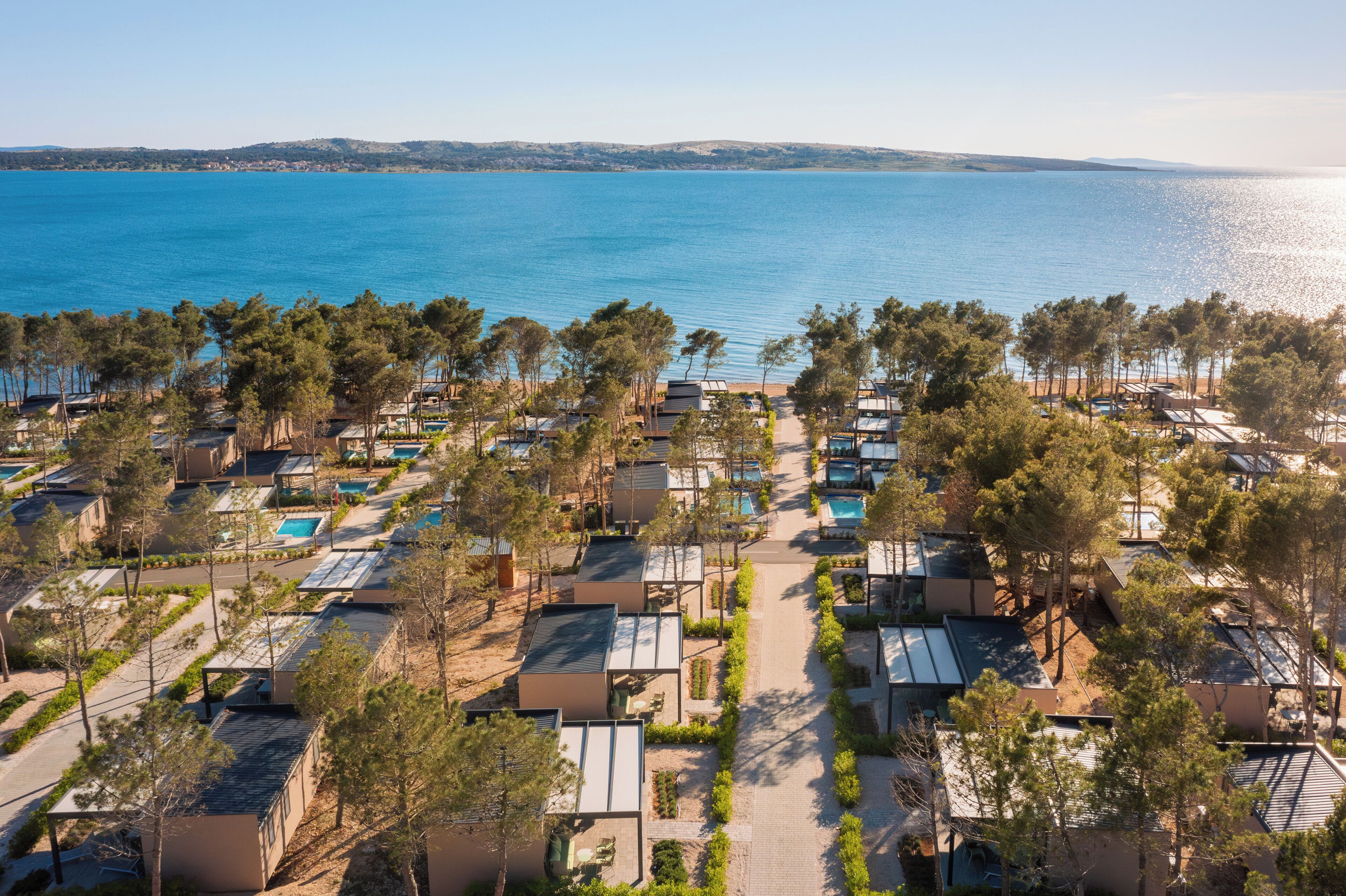 A coastal resort with multiple bungalow-style accommodations, each with a private pool, surrounded by pine trees and overlooking the sea.