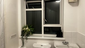 Baño con inodoro, bidé y lavamanos. Hay una ventana con cortina y una planta en el alféizar.