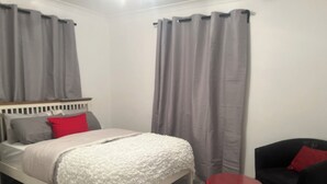 Un dormitorio con una cama, cortinas grises, una almohada roja, una silla negra y una mesa redonda negra.