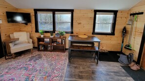 A cozy room with a sofa, a small table with a plant, a TV mounted on the wall, and a window with a view of trees. - Travelers Haven (Whitefield)
