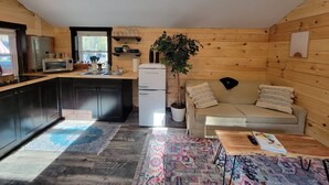 A compact kitchen with a refrigerator, microwave, and sink. There is a beige sofa with cushions and a small table with a book. A potted plant is near the sofa. - Travelers Haven (Whitefield)