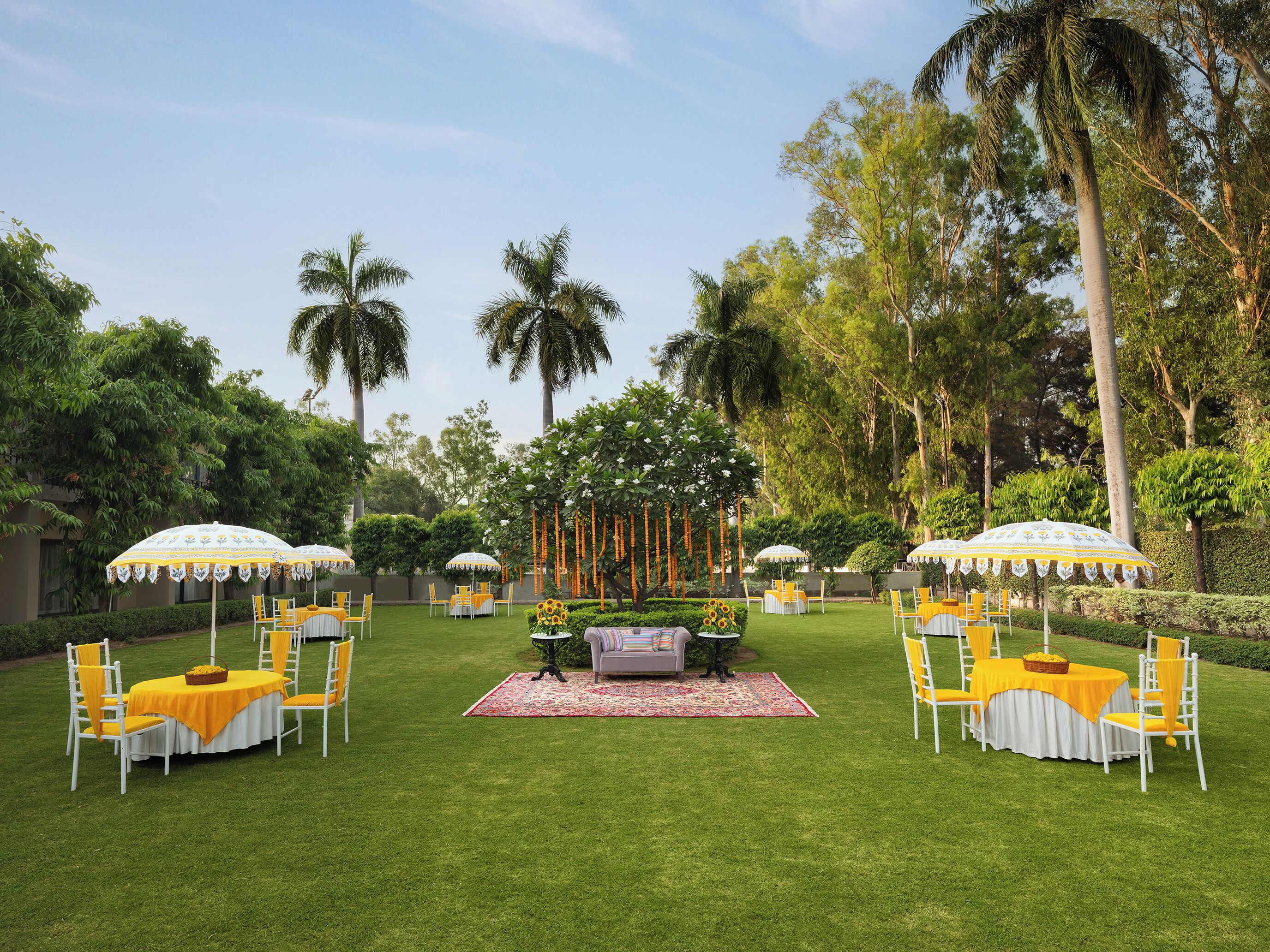 An outdoor garden setting with multiple round tables and chairs, surrounded by lush greenery and palm trees.