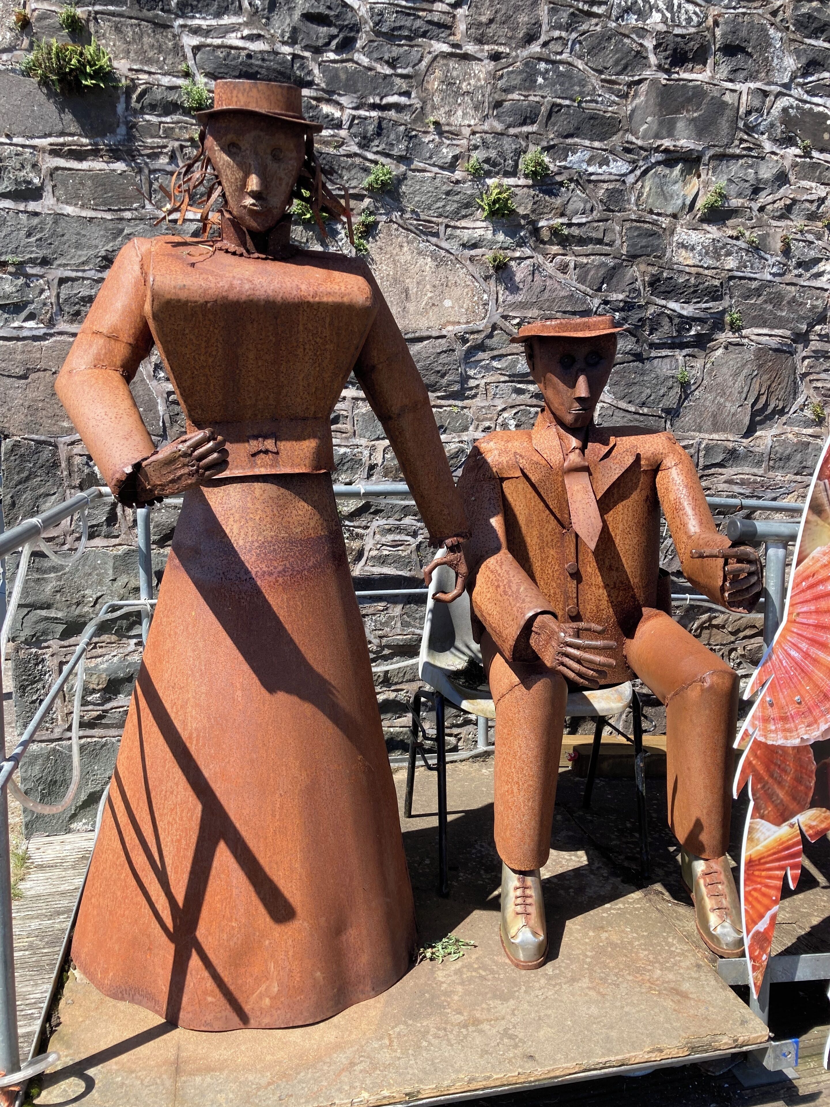 A sculpture of a woman and a man in rust-colored attire, seated on a platform with a stone wall background.