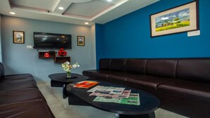 A living room with a brown leather sofa, a black coffee table with magazines, a TV on a stand, and a framed picture on the wall. - MeroStay 127 Divine Guest House (Kathmandu)