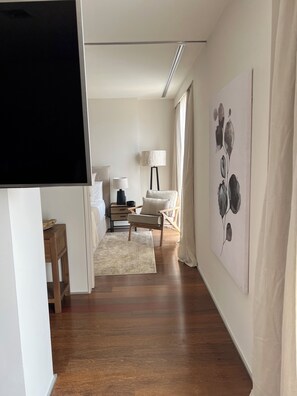 A hotel room with a wooden floor, a bed with white linens, a nightstand with a lamp, a chair, and a painting on the wall.