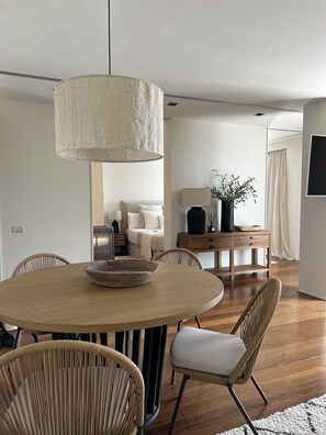 A dining area with a round wooden table, wicker chairs, a large hanging light fixture, and a wooden sideboard with a plant.