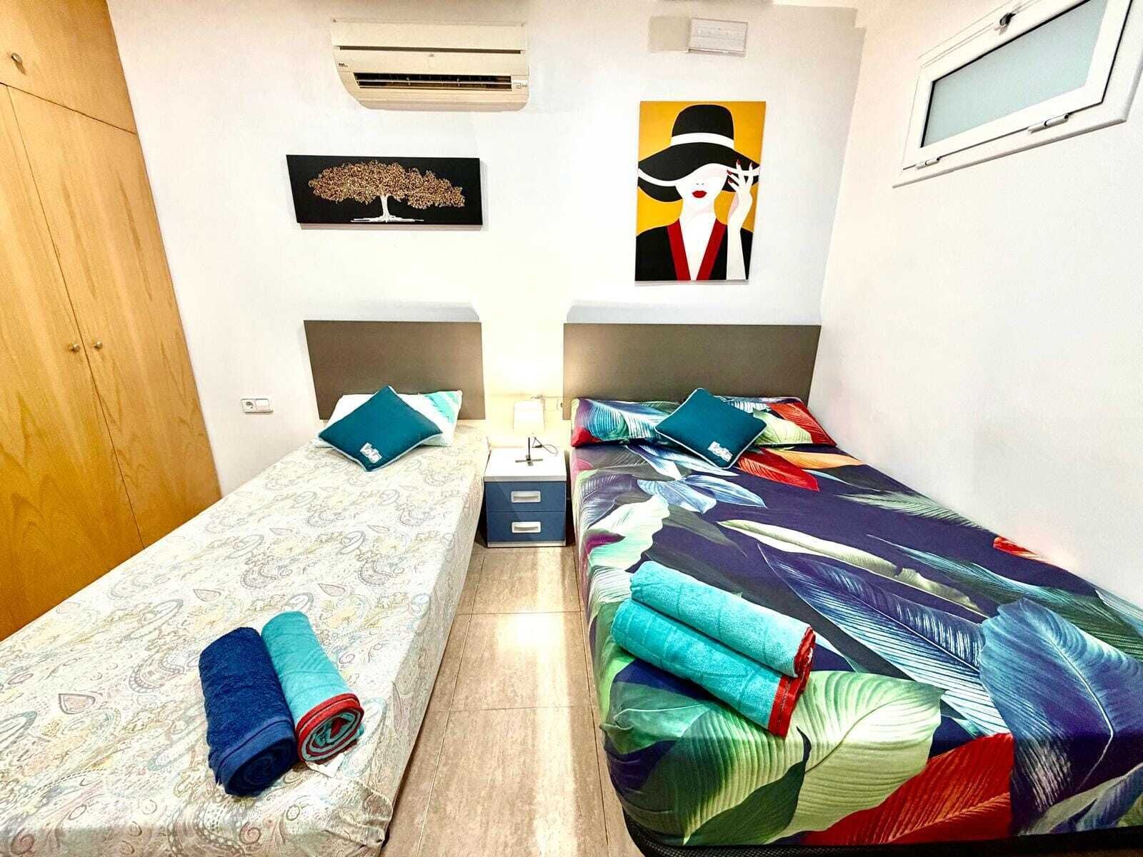 Una habitación pequeña y bien iluminada con dos camas, un armario, un equipo de aire acondicionado y obras de arte en las paredes.