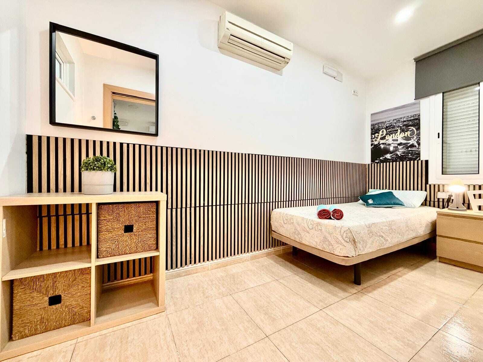 Un dormitorio moderno con cama, mesita de noche, lámpara, equipo de aire acondicionado, espejo y una pared con listones verticales.