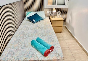 A single bed with a patterned bedspread, a blue pillow, and a rolled-up towel with red trim. There is a bedside table with a lamp and a letter 'B' decoration.