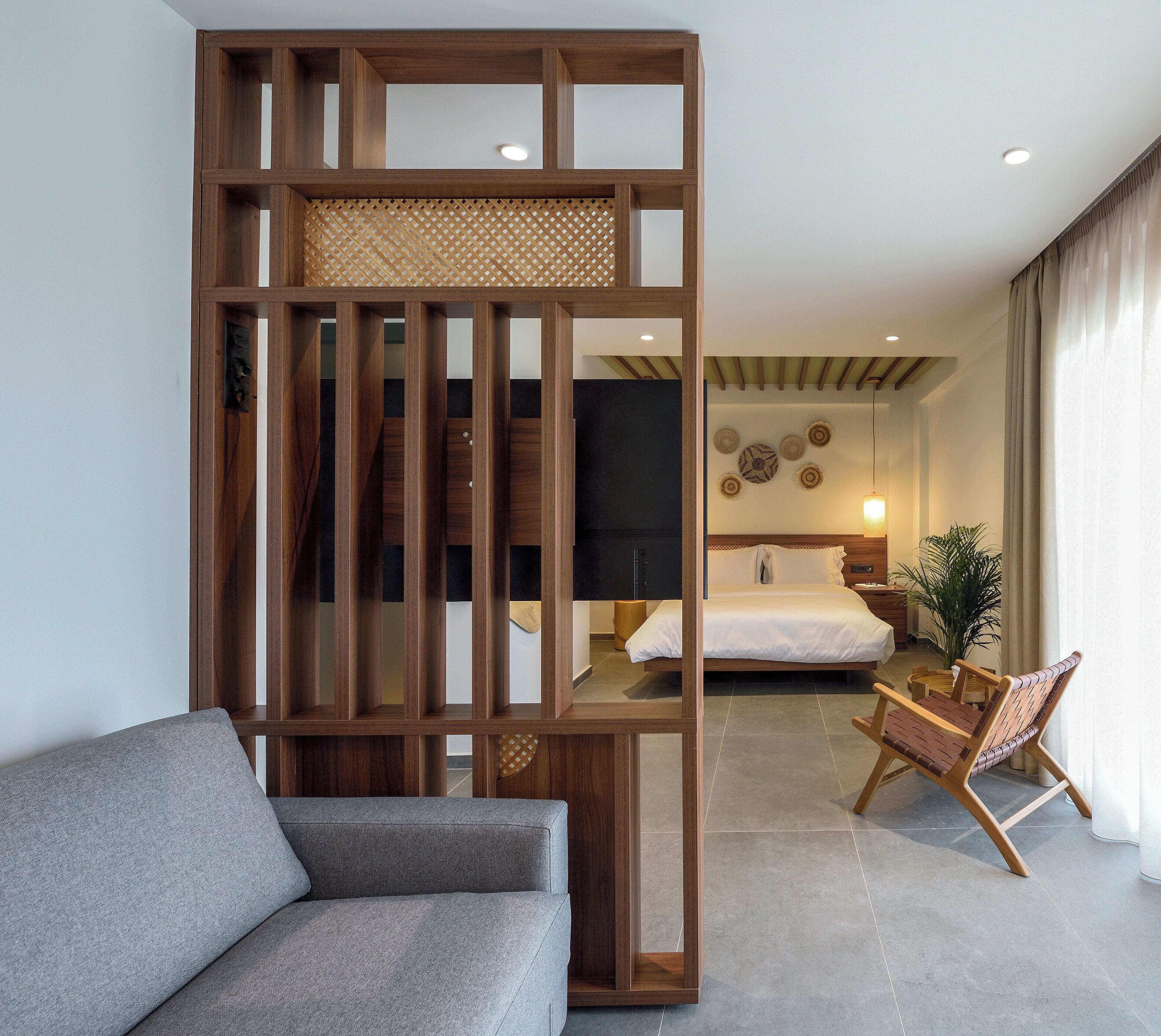 A modern hotel room with a wooden partition, a grey sofa, a bed with white bedding, a wooden nightstand, and a potted plant.
